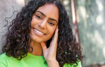 Young woman with curly hair smiling at the camera, wearing a bright green shirt and touching her face with her hand.