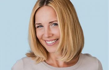 A smiling woman with short blonde hair wearing a beige top against a light blue background.