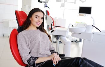 A woman sitting in a red dental chair in a modern dental office, smiling at the camera.