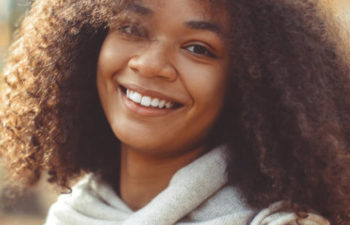 A young woman with curly hair smiling outdoors on an autumn day, wearing a scarf and plaid jacket.