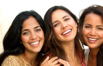 Three women smiling at the camera, standing close together, with a softly lit background.